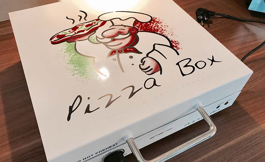 Pizzabox vpn Emerio auf Küchentisch - weißer Aluminiumkasten zum Öffnen mit Aufdruck Pizzabox und ein Cartoon Pizzabäcker
