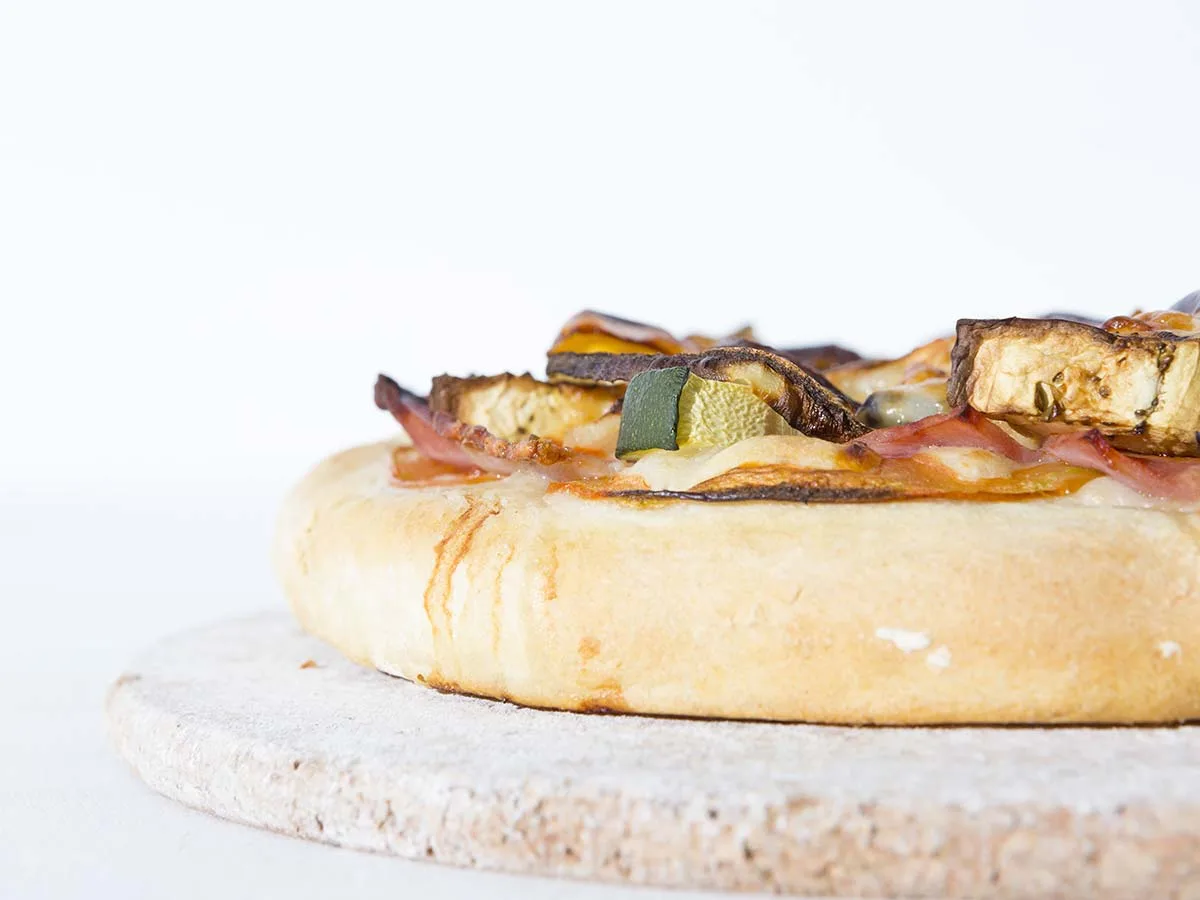 Fertig gebackene Pizza mit Zucchini, Aubergine und Schinken auf einem runden Pizzastein, Nahaufnahme der goldbraunen Kruste
