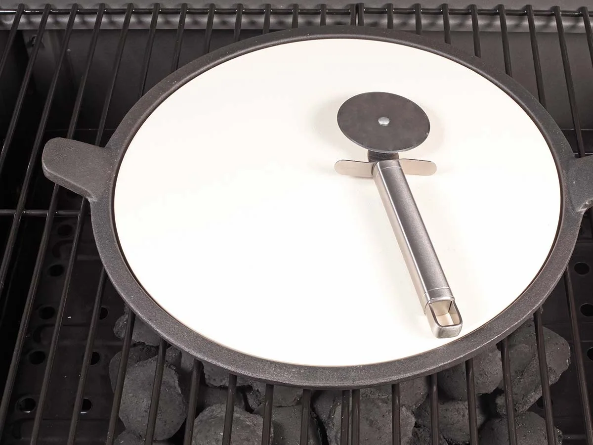 Runder heller Pizzastein auf Metallgestell mit zwei Griffbügeln, darauf ein Pizzaroller aus Edelstahl