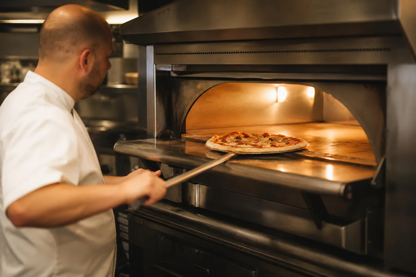Pizzabäcker schiebt eine Pizza in einen großen Edelstahl-Pizzaofen in einer Gastronomieküche