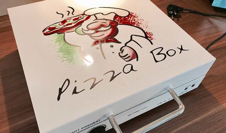 Pizzabox vpn Emerio auf Küchentisch - weißer Aluminiumkasten zum Öffnen mit Aufdruck Pizzabox und ein Cartoon Pizzabäcker